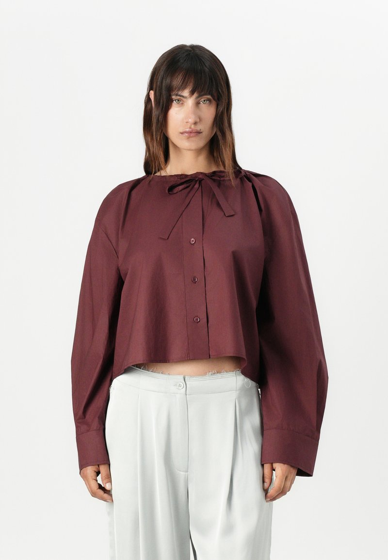 Burgundy cropped blouse with a tie neckline, button front, and long sleeves; worn with light grey high-waisted trousers. Smooth texture.