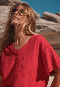 T-shirt en coton rouge avec un col en V, des manches courtes et une poche avant ; texture légère, coupe décontractée, sur un fond rocheux.