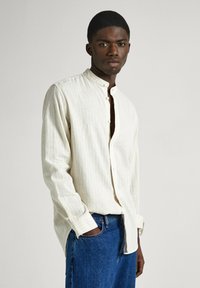Camicia a maniche lunghe di un beige chiaro con colletto alla coreana, caratterizzata da sottili righe verticali e una vestibilità rilassata, abbinata a jeans blu scuri.