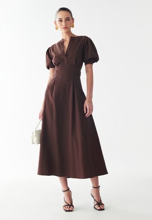 Ruda midi suknelė su pūstuvais rankovėmis ir giliu V formos iškirpimu. Apatinė dalis dekoruota klostelėmis, o juosmuo prigludęs. Aksesuarai: juodi sandalai su dirželiais ir balta rankinė.