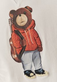 Hímzett maci, narancssárga bolyhos kapucnis pulóvert, kék farmert és fekete, fehér részletekkel díszített sportcipőt visel, fehér textil háttéren.