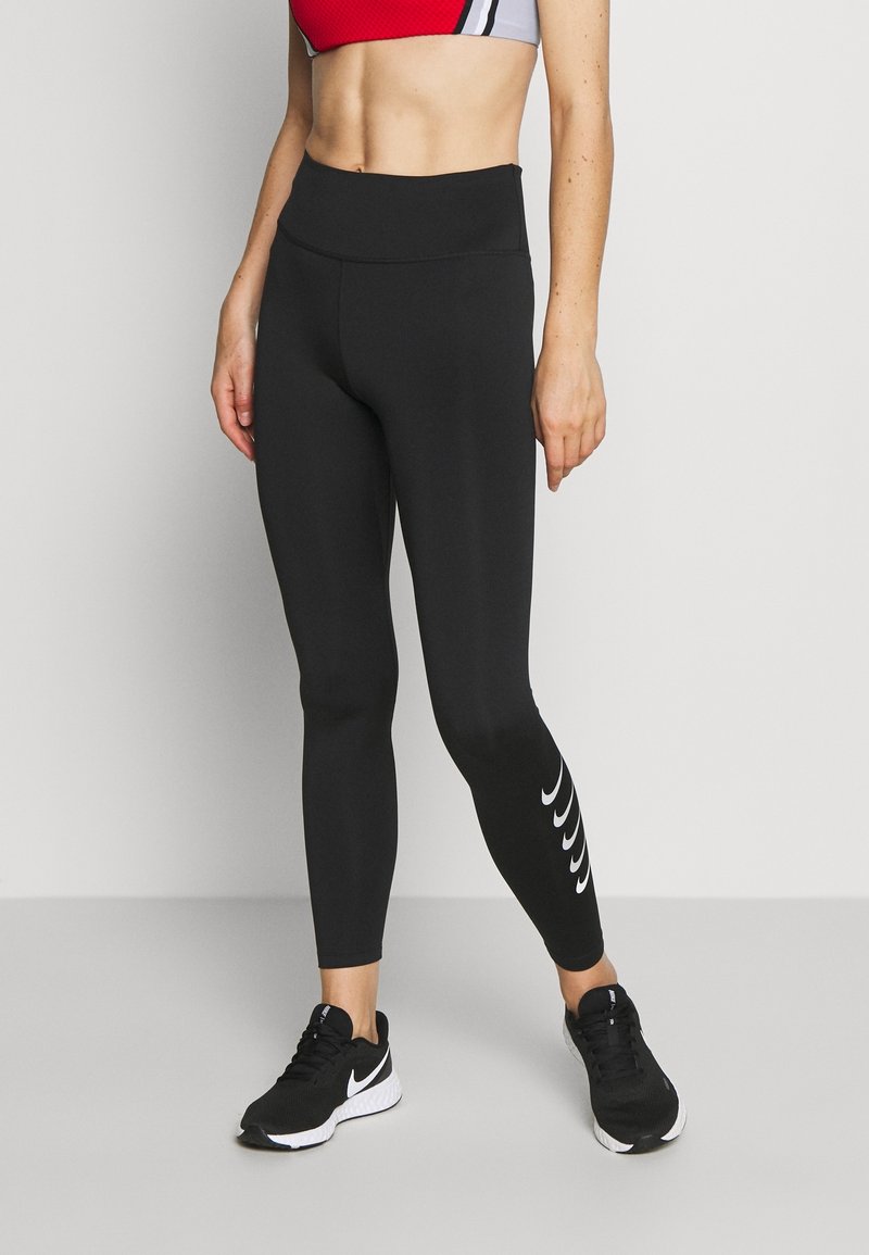 Leggings negras de material elástico con cintura alta. Presenta acentos en forma de ola blanca en la parte inferior de la pierna. Combinados con zapatillas deportivas negras.