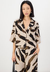 Camicetta beige a maniche corte con motivo a strisce di tigre in nero e marrone. Presenta un colletto, una tasca e una vestibilità ampia e rilassata.