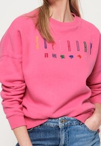 Pink sweatshirt with long sleeves, featuring a multicolored "SUPERDRY" logo across the chest, made from soft, textured fabric.