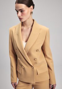 Béžový dvouřadý blazer se zlatými knoflíky, s klopami a předními kapsami. Hladká látka a strukturovaný střih.