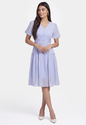 Frau mit langen dunklen Haaren, die ein helllila knielanges Kleid mit kurzen Ärmeln und weißen Absätzen trägt, steht vor einem einfarbigen Hintergrund.