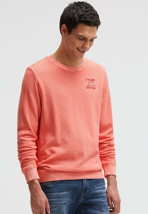 Mann trägt korallenfarbenen Langarm-Sweatshirt mit weißem Text auf der Brust und blaue Jeans, schaut nach unten und lächelt vor einfarbigem Hintergrund.