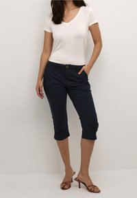 Námořnické zkrácené kalhoty s knoflíkovým zapínáním, předními kapsami a hladkým povrchem, spárované s přiléhavým bílým tričkem s výstřihem do V a hnědými sandály na podpatku.