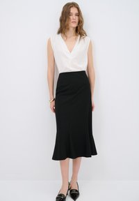 Valkoinen ilmanhihoilla oleva toppi V-päällä ja kultatehosteilla, yhdistetty mustaan liehuvaan hameeseen. Mustat teräväkärkiset kengät täydentävät asua.