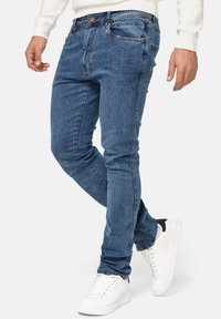 Jean en denim bleu à coupe slim, doté de cinq poches, d'un détail de whiskering subtil et associé à des baskets blanches.
