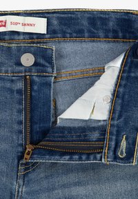 Vaqueros de mezclilla azul oscuro con un ajuste skinny, que presentan una bragueta con cremallera, herrajes en tono cobre y detalles de costura amarilla en contraste.