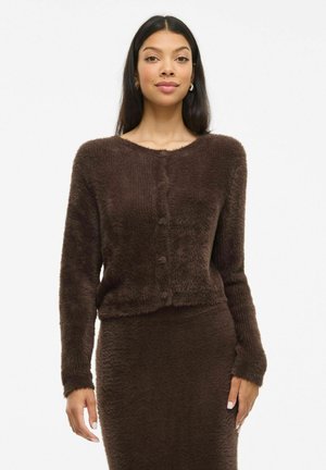Cardigan court en peluche marron avec un col rond, fermeture à boutons et manches longues. La texture est douce et moelleuse, s'harmonisant avec une jupe assortie.