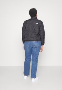 Svart isolerad jacka med hög krage, texturerad yta och logotyp på övre ryggen, kombinerad med ljusblå jeans och vita skor.