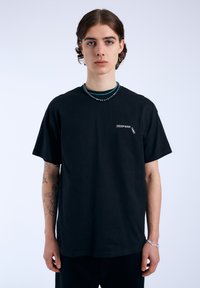 Camiseta de algodón negra con mangas cortas, cuello redondo y pequeño texto gráfico blanco en el lado izquierdo del pecho. El modelo lleva collares de cuentas superpuestos.