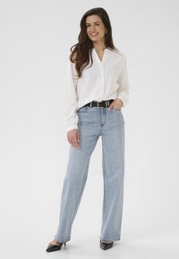 Lächelnde Frau mit langen Haaren, die ein weißes Hemd mit Knöpfen, hellblaue Jeans, einen schwarzen Gürtel und schwarze spitze Schuhe trägt und vor einem weißen Hintergrund steht.