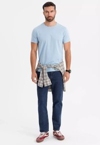 Světle modré bavlněné tričko, modré džínové kalhoty, károvaná flanelová košile uvázaná v pase, červené a bílé tenisky, jednobarevné pozadí.