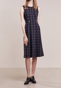 Marinblå klänning med vit rutmönster, ärmlös design, A-linjär silhuett, med dekorerade knappar och sidfickor. Bärs med svarta skor.
