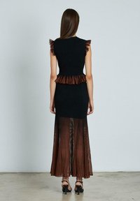Mujer vestida con un vestido negro sin mangas, con hombros y cintura con volantes, que presenta una falda marrón sheer con pliegues, de pie sobre un suelo de hormigón.