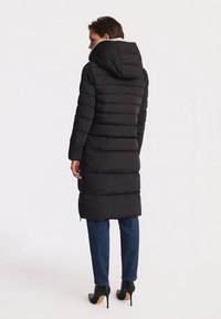 Czarna pikowana kurtka puchowa z kapturem, z poziomym przeszyciem, o długości do kolan, z ozdobami po bokach na zamek. Noszona na ciemnych dżinsach.