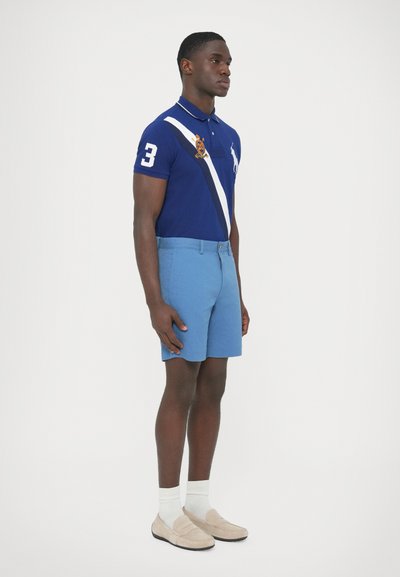 Modré polo s bielym pruhom a číslom 3, v kombinácii s svetlomodrými šortkami a béžovými mokasínami, má texturovaný povrch a jemné značkové prvky.