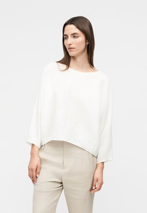 Femme aux cheveux bruns et raides portant un haut blanc à manches longues ample et un pantalon beige taille haute, sur un fond clair uni.