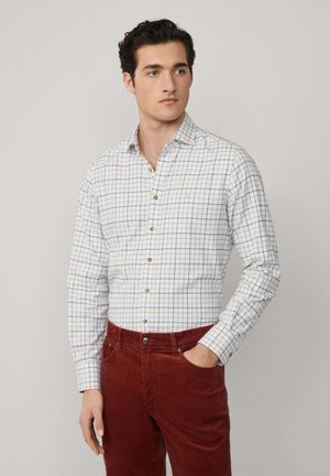 Camisa blanca a cuadros con rayas azules y naranjas, de mangas largas, cuello abotonado, combinada con pantalones de pana rojos, de pie contra un fondo claro.