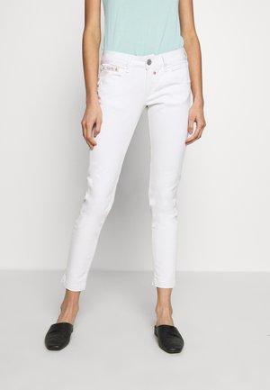 Kvinde iført tætsiddende hvide jeans og sorte slip-on sko, stående mod en ensartet lys baggrund.