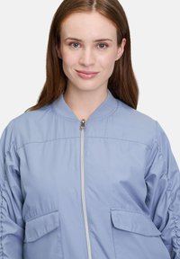 Hellblau Bomberjacke mit Frontreißverschluss, geripptem Kragen und zwei Brusttaschen. Der Stoff hat eine glatte Oberfläche und einen leicht gepolsterten Look.