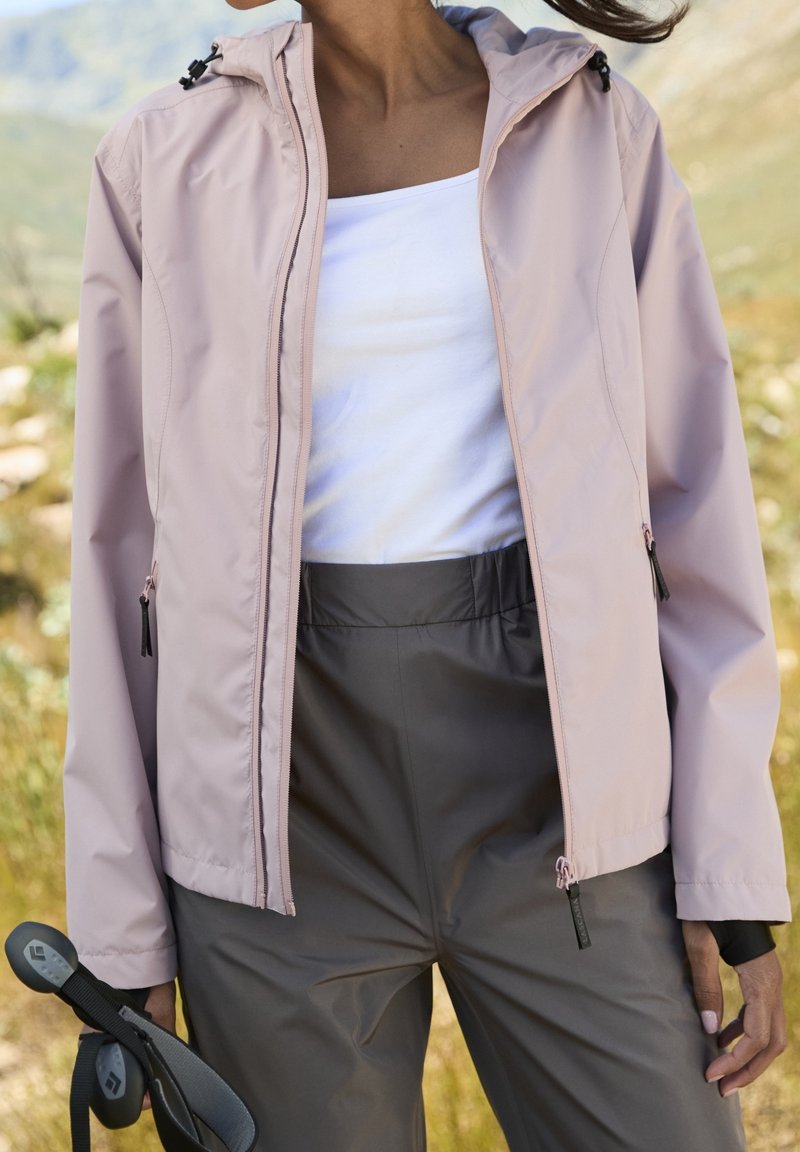 Femme à l'extérieur portant une veste rose clair, un haut blanc et un pantalon gris, tenant des bâtons de randonnée avec un paysage naturel en arrière-plan.