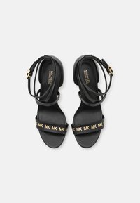 Czarne płaskie sandały z paskami na kostki, złotym zapięciem i złotymi logo "MK" na przednich paskach, markowe wkładki Michael Kors.