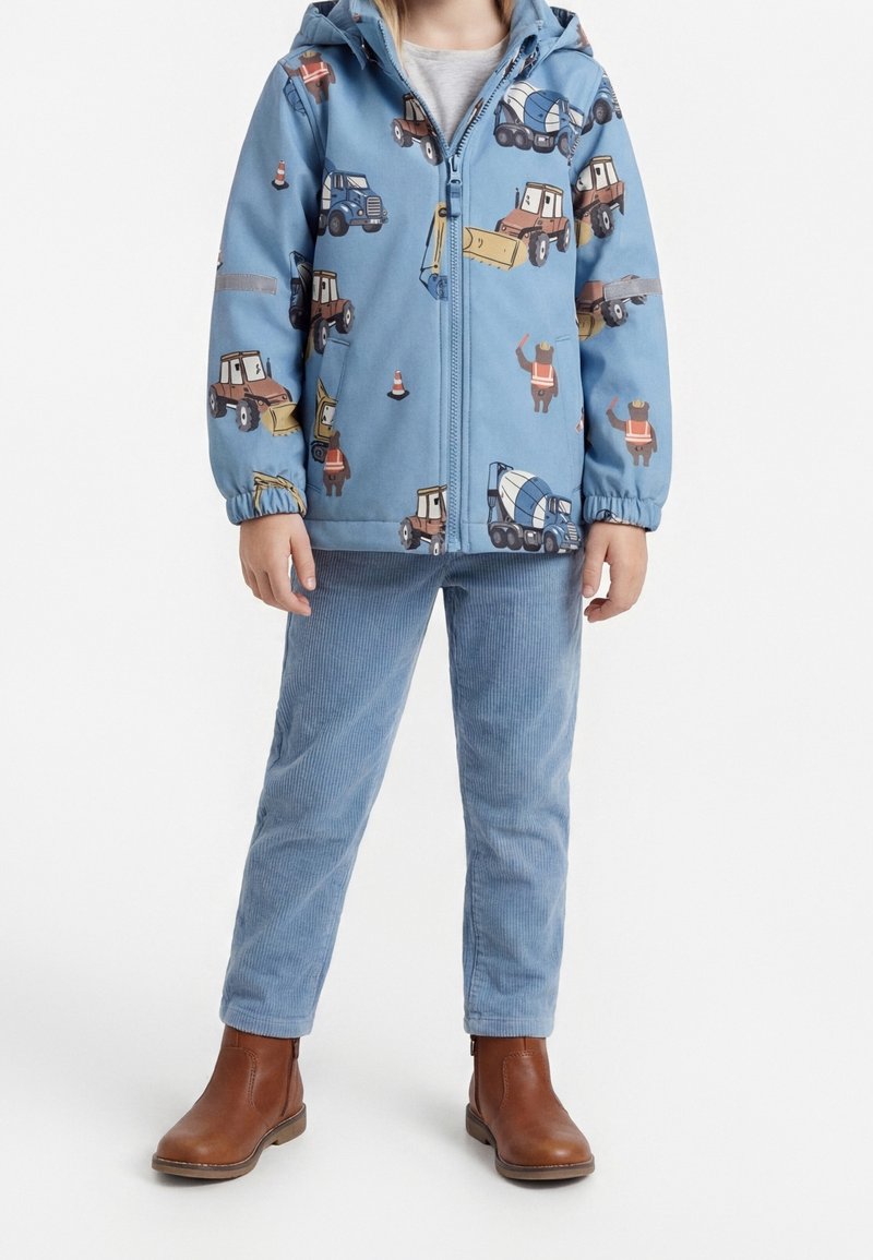 Enfant portant une veste à capuche bleue avec des motifs de véhicules de chantier et d'ouvriers, pantalon bleu clair et bottines marron.