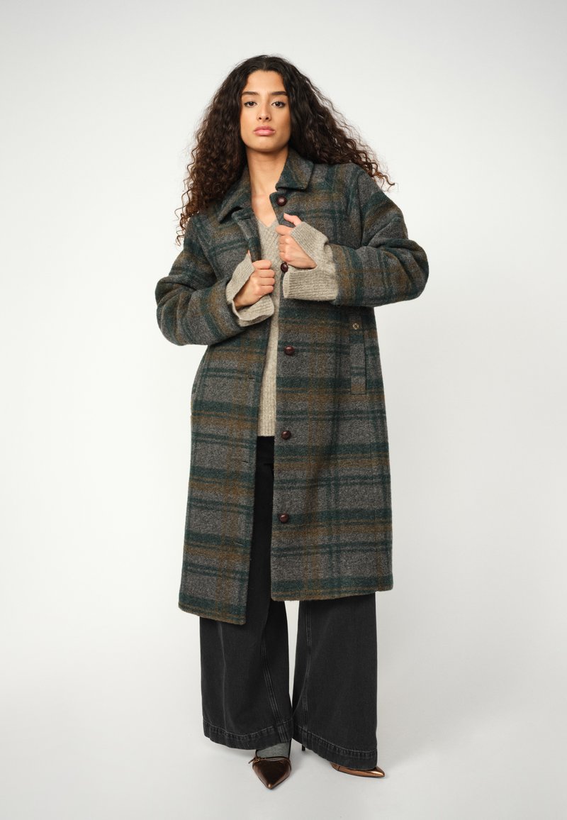 Karierter Wollmantel in Grau, Grün und Braun mit Kragen, Knopfleiste und aufgesetzten Taschen. Kombiniert mit einem beigen Pullover und einer weiten schwarzen Hose.
