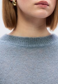 Ανοιχτό μπλε πλεκτό πουλόβερ με στρογγυλή λαιμόκοψη. Ορατή απαλή υφή, με διακριτικό σχέδιο. Χρυσά σκουλαρίκια σε σχήμα σταγόν ας εμφανή.