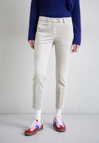 Person, der beigefarbene, gekürzte Hosen, weiße Socken und rote, rosa sowie blaue Turnschuhe trägt und vor einer schlichten hellgrauen Wand steht.