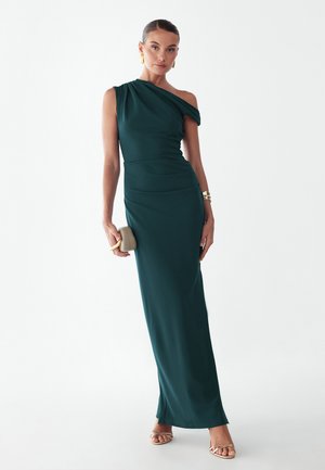 Kobieta w ciemnozielonej, długiej do ziemi sukni na jedno ramię, trzyma beżową kopertówkę, nosi złote kolczyki, bransoletkę oraz beżowe sandały na paski.