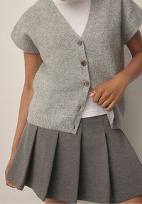Femme portant un gilet en maille gris à boutons sur une chemise blanche, associé à une jupe plissée grise, ajustant le gilet avec sa main.
