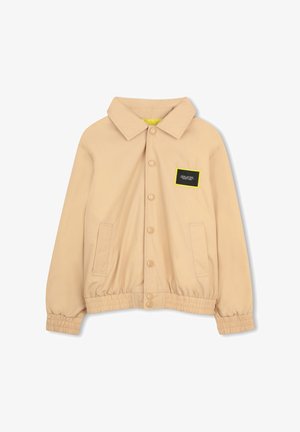 Beige jas met een kraag, drukknoopsluiting, elastische zoom en twee zijzakken. Heeft een zwart label met een gele omlijning.