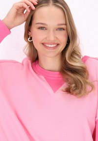 Felpa over-size rosa con scollo a V, con una finitura testurizzata. Il modello indossa orecchini a forma di cuore bianchi e ha i capelli mossi.