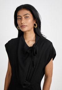 Robe noire avec un tissu texturé, présentant un détail de col noué et des manches courtes et larges. Des boucles d'oreilles en or ajoutent du contraste.