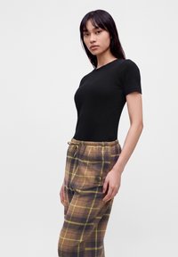 Jeune femme aux longs cheveux foncés portant un top noir à manches courtes et un pantalon à carreaux marron avec cordon, debout devant un fond uni.