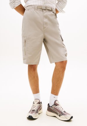 Άνδρας που φοράει μπεζ cargo σορτς, λευκές κάλτσες και αθλητικά παπούτσια σε μπεζ και καφέ αποχρώσεις, στέκεται με τα χέρια πίσω από την πλάτη.