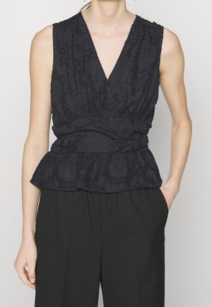 Czarna teksturowana bluzka bez rękawów z dekoltem w serek, talią marszczoną i falbankowym pepłum, noszona z czarnymi spodniami z wysokim stanem, jedna ręka w kieszeni.