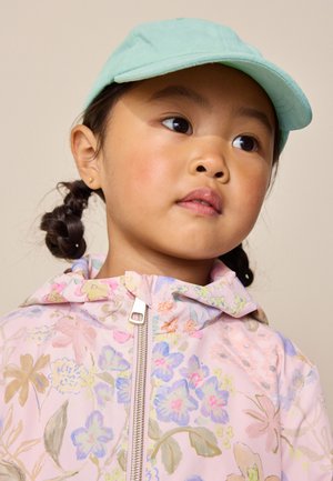 Ragazza giovane con capelli intrecciati che indossa un berretto verde menta e una giacca rosa floreale con zip, guardando leggermente di lato.