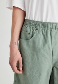 Pantaloni scurți verzi din in, cu o talie elastică, având o buzunar lateral cu un mic nasture auriu. Textura pare moale și ușoară.