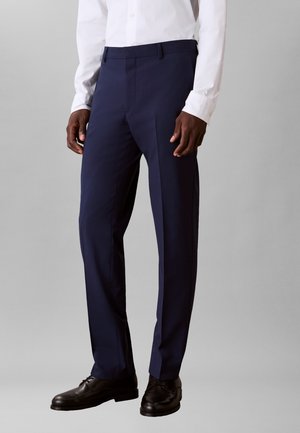 Mann trägt marineblaue, eng anliegende Anzughose, schwarze Lederschuhe und ein weißes, geknöpftes Hemd und steht vor einem schlichten grauen Hintergrund.