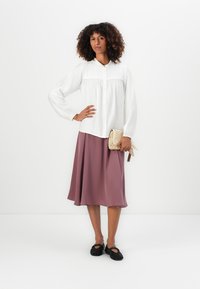 Kvinne med krøllete hår iført en hvit bluse og mauve midi-skjørt, som holder en vevd clutch, står med den ene hånden på hoften og svarte sko.