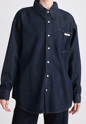 Personne portant une chemise en denim bleu foncé à boutons, manches longues et une poche poitrine étiquetée Calvin Klein.