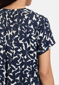 Blusa blu navy con motivo floreale bianco, maniche corte, scollatura plissettata, tessuto morbido e vestibilità comoda, che mette in risalto un design delicato.
