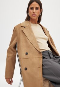 Beige nubukist topeltčine coat, millel on suured kraed ja mustad nupud, kantud heleda sviitri ja hallide pükste üle. Sile tekstuur, mugav lõige.