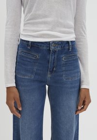 Jean en denim bleu avec une coupe droite, deux poches arrière et une texture légère, légèrement délavée. Porté avec un top blanc à manches longues.